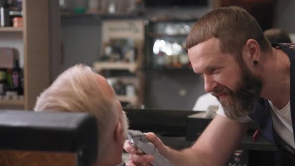 Barbiere alla moda utilizza forbici e un pettine per tagliare i capelli a un uomo più anziano in una sedia parrucchieri in un salone da uomo — Video Stock