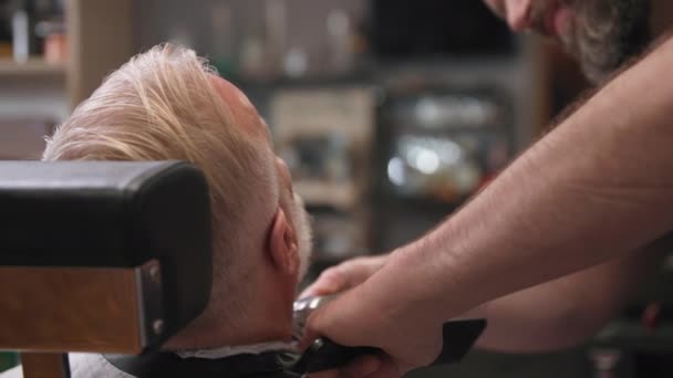 Berber dükkanı, profesyonel berber, kuaför koltuğunda yaşlı stil sahibi bir müşteri için saç kesiciyle sakal ve tarak kesiyor. — Stok video