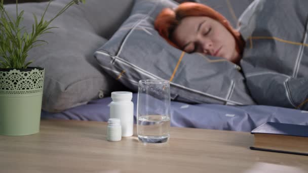 Egészségügyi ellátás, orvosi gyógyszerek egy pohár víz stand az éjjeliszekrényen háttér egy alvó nő az ágyban, közelkép — Stock videók