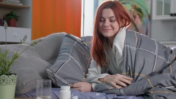 Szczęśliwa kobieta bierze tabletki lub witaminy i pije je je z chłodną czystą wodą po przebudzeniu ze snu leżącego w łóżku w pokoju, uśmiechając się i patrząc w kamerę — Wideo stockowe