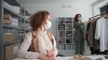 Çevrimiçi dükkan, tıbbi maskeli genç profesyonel kadın satıcılar Coronavirus 'un butik deposunda oturması nedeniyle bilgisayardan alışveriş yapanlarla uzaktan haberleşiyorlar.