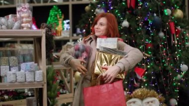 Tatil alışverişi yapan, elinde hediyeler olan genç ve çekici bir kadın, alışveriş merkezindeki bir mağazada satışta aile için raflarda bulunan ürünleri seçiyor.