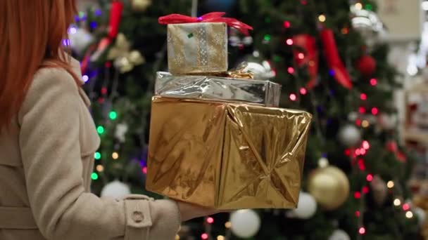 Voorbereiding op de wintervakantie, een vrouw met geschenken in haar handen op de achtergrond van een kerstboom versierd met gekleurde bloemenslingers in een winkel — Stockvideo
