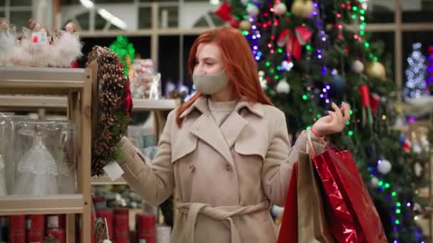 Virüs ve enfeksiyondan korunmak için koruyucu maskeli genç bir kadın tatil için evi dekore etmek üzere yeni yıl süslemelerini seçiyor. — Stok video