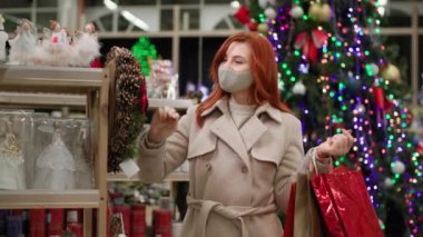 Koruyucu maskeli genç bir kadın kış tatilinde ailesine hediye alıyor ve süpermarkette yeni yıl dekoruyla başparmak kaldırıyor.