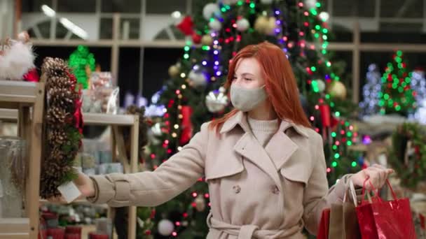 Mooie jonge vrouw in een beschermend masker kiest geschenken voor haar familie op een nieuwjaarsvakantie in een winkelcentrum — Stockvideo