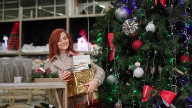 Mutlu kadın, yeni yıl tatillerinde aile alışverişinde mutlu, gülümseyen ve kameraya bakan alışveriş merkezindeki Noel ağacının arka planındaki şenlik kutusunu gösteriyor.