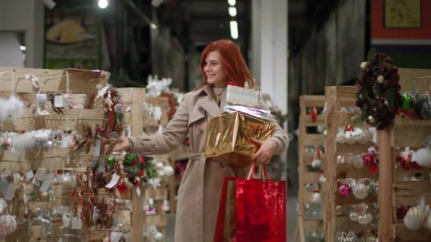 Kış tatili için hazırlanan, elinde hediyeler olan çekici bir kadın süpermarkette dekorasyon seçiyor, gülümsüyor ve kameraya bakıyor ve sınıfını gösteriyor. — Stok video