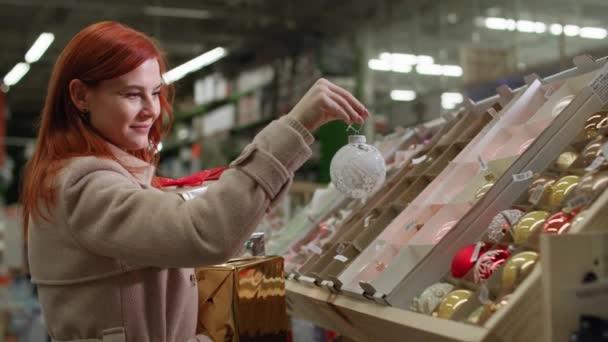 Молодая женщина выбирает новогодние игрушки для украшения елки на зимние праздники в розничном магазине — стоковое видео