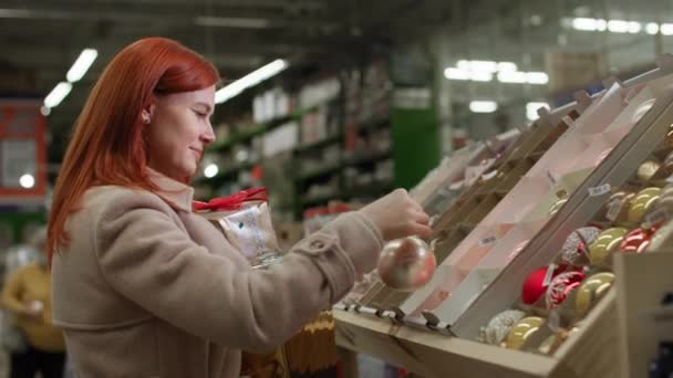 Charmante vrouwelijke shopper kiest nieuwjaar speelgoed te versieren kerstboom voor de wintervakantie in het winkelcentrum, glimlacht en kijkt naar de camera — Stockvideo