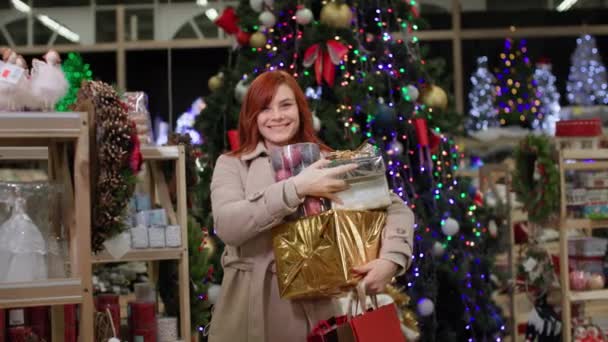 Nový rok prodej, portrét radostné ženy s dárky v rukou u polic se zbožím na pozadí vánočního stromečku s věnci, s úsměvem a pohledem do kamery — Stock video