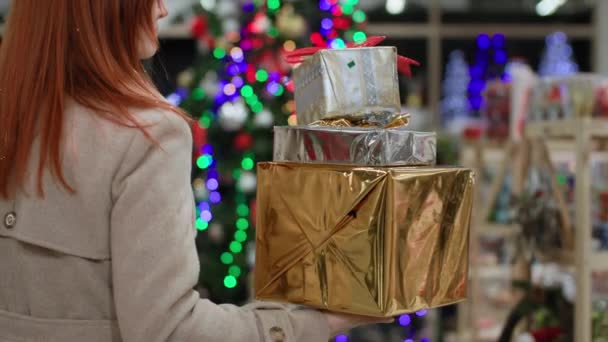 Voorbereiding op de feestdagen, een jonge vrouw houdt mooie geschenken in haar handen tegen de achtergrond van veelkleurige lichten in de winkel, een close-up plan — Stockvideo