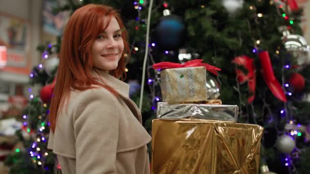 Retrato de mujer hermosa con regalos en las manos sobre el fondo del árbol de Navidad con luces en el supermercado, sonriendo y mirando a la cámara — Vídeos de Stock