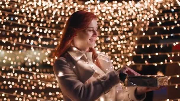 Рождественский вечер, очаровательная женщина радуется открытию поздравительной коробки с подарком на фоне огней в парке, улыбаясь и глядя в камеру — стоковое видео