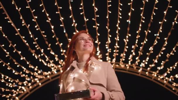 Charmante vrouw in bril met cadeau in haar handen wandelingen in prachtig park versierd met een boog van bloemenslingers op kerstavond, glimlachen en kijken naar de camera — Stockvideo