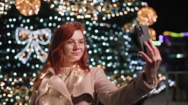 Yaşam tarzı, gözlüklü ve şapkalı hoş bir kadın, gece geç saatlerde bir şenlik ağacının özçekimini yapıyor ve parkta yeni yıl çelenkleri yapıyor.