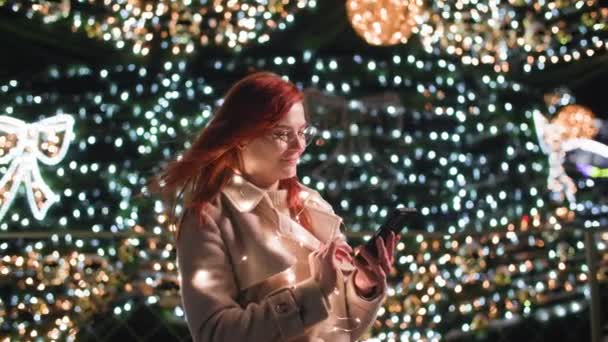 Mulher encantadora com um telefone celular escreve parabéns SMS para parentes e amigos na véspera de Ano Novo perto de uma árvore de Natal decorada com luzes — Vídeo de Stock