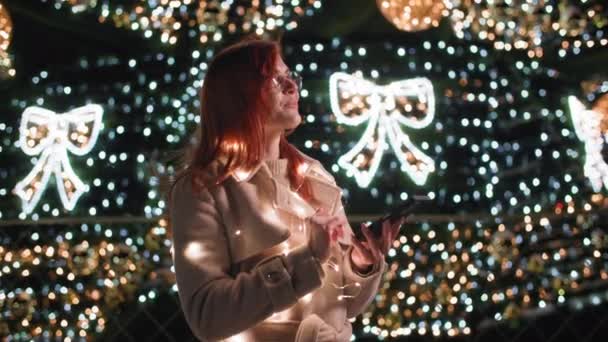 Mladá žena v brýlích s mobilním telefonem chatování s přáteli stojící na pozadí vánočních stromků zdobené věnce v parku pozdě večer — Stock video