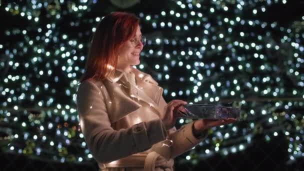 Portré egy boldog nő örömmel megnyitja szilveszteri ajándékát, örömmel néz a kamerába, és mosolyog háttér karácsonyfa díszített koszorú — Stock videók