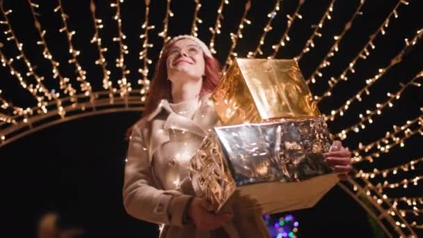 Świąteczna atmosfera, radosna kobieta spaceruje z prezentami w rękach dla rodziny i lokalnie patrzy na łuk świecących świateł z girlandy podczas późnego wieczornego spaceru po mieście — Wideo stockowe