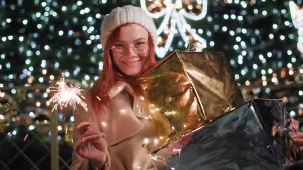 Feestelijke sfeer, portret van lachende vrouw in hoed en bril met geschenken in de hand en met bingal lichten staat achtergrond van kerstboom en lichten 's nachts in het park — Stockvideo