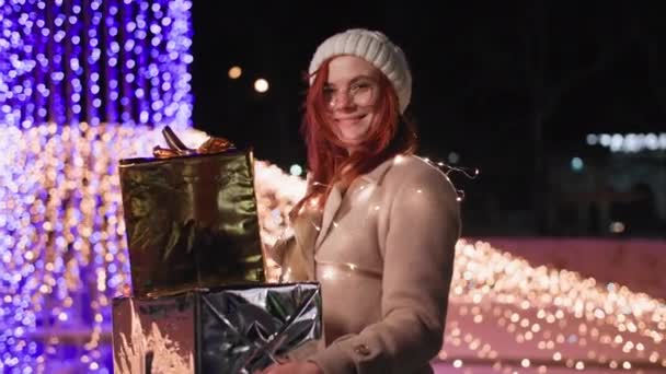 Újévi hangulat, bájos nő kalapban és szemüvegben ajándékokkal a kezében áll egy éjszakai parkban díszített színes fények, mosolyog, és néz a kamerába — Stock videók