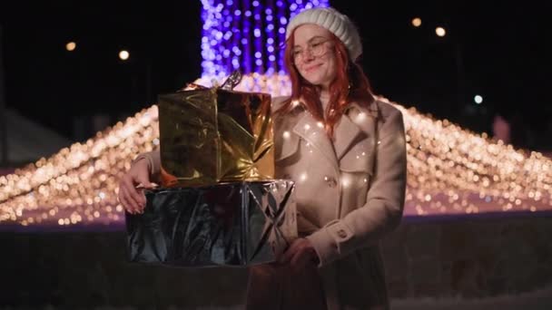 Портрет молодой женщины в шляпе стоит с подарками в руках на фоне мерцающих огней в городе в канун Нового года — стоковое видео