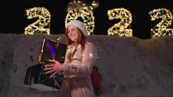 Téli ünnepek, nő szemüvegben ajándékokkal forog a háttér szilveszteri fények a parkban — Stock videók