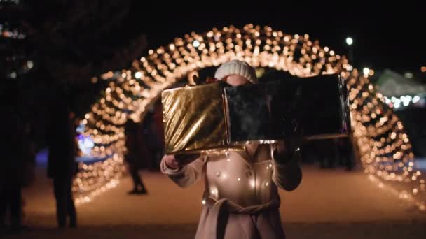 Зимние праздники, улыбающаяся женщина в очках с подарками в руках стоит на фоне ярких огней, улыбается и смотрит в камеру — стоковое видео