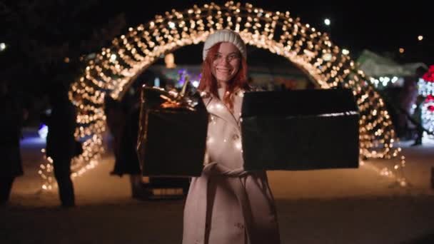 Portrait of a joyful female with gift boxes in her hands against the backdrop of an arch of glowing light bulbs in a night park — Stock Video