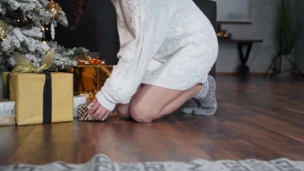 Férias de inverno, uma mulher encantadora caminha sobre os dedos dos pés para um presente sob a árvore de Natal, leva em suas mãos, sorri e olha para a câmera — Vídeo de Stock
