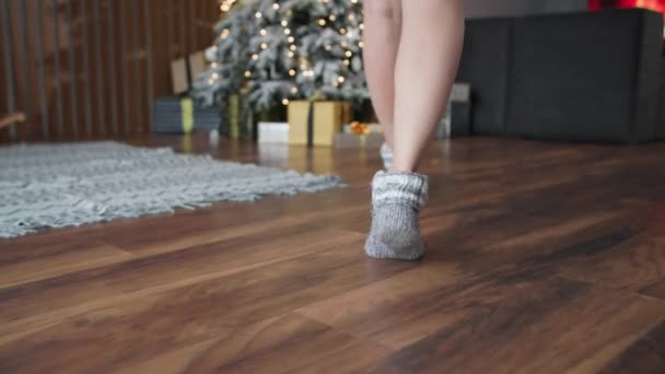 Jonge vrouw loopt op tenen naar de kerstboom voor een geschenk, neemt het in haar handen terwijl zitten in een gezellige kamer op een winteravond — Stockvideo