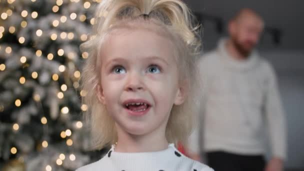 Porträt eines charmanten süßen Mädchens, das lächelt und sich im Hintergrund des Neujahrsbaums umsieht — Stockvideo