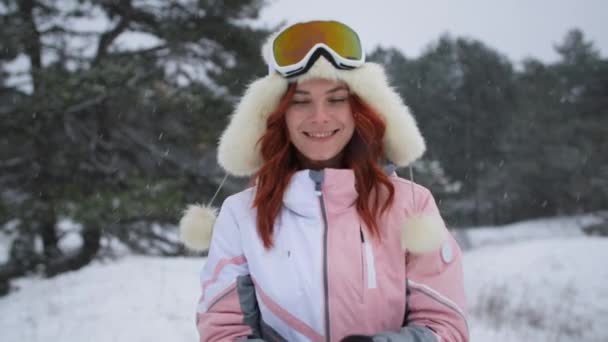 Hermosa mujer sonriente en sombrero y gafas de pie en el bosque en el día de invierno nevado al aire libre, chica levanta las manos y mira hacia arriba en el soplado de copos de nieve directamente a la cámara — Vídeos de Stock