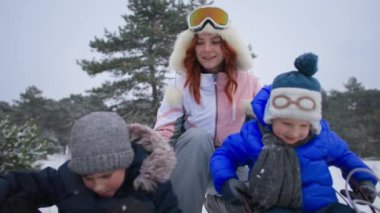 eğlenceli kış tatilleri, kızaktaki anne ve oğulları ormanda eğleniyor, çocuklar açık havada tepeden kayıyor.