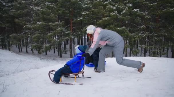 冬の森の親を持つ子供たち屋外活動中に子供と一緒に丘から雪とそりを楽しむ母親 — ストック動画