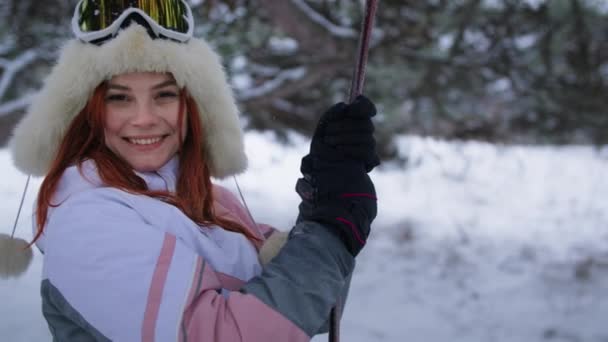 Joven mujer disfruta de la naturaleza de invierno y mira a la cámara, chica feliz en sombrero caliente y guantes paseos en un columpio en el bosque — Vídeo de stock