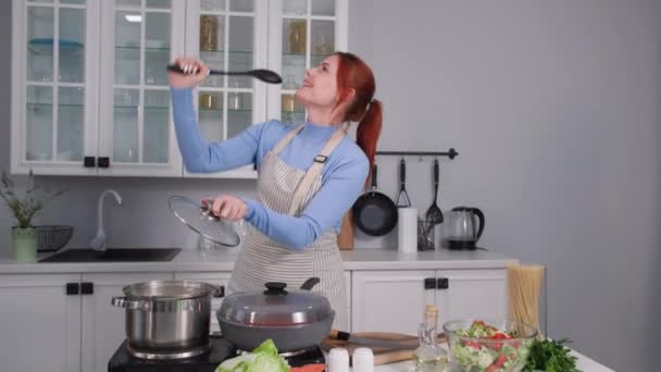 Adorable ama de casa divirtiéndose bailando y cantando usando una cuchara como micrófono mientras está de pie en la cocina mientras cocina — Vídeos de Stock
