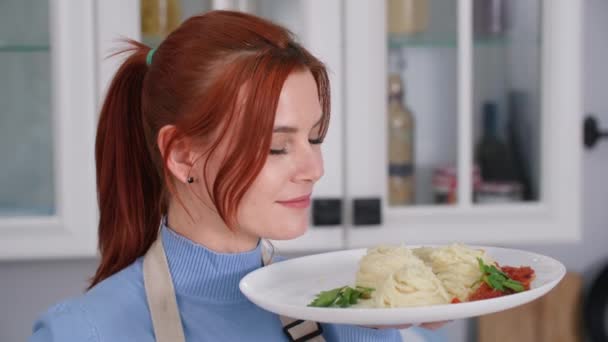 Portré aranyos fiatal nő tányér spagetti a kezében áll a háttérben a konyha, mosolygós és a kamerába néz — Stock videók