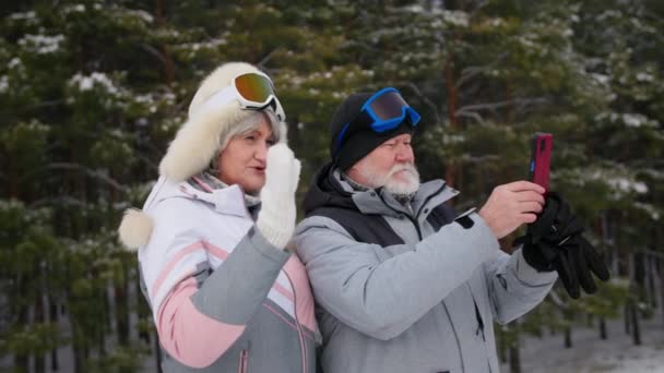 Modern idős pár mobiltelefonnal forgatás téli erdő, ősz hajú nyugdíjasok meleg ruhában és síszemüveg együtt töltik az időt a szabadban — Stock videók