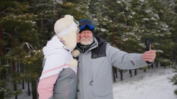 Glad modern äldre par tar foto på mobiltelefon på naturen bakgrund, mormor kysser morfar i vinterskogen — Stockvideo