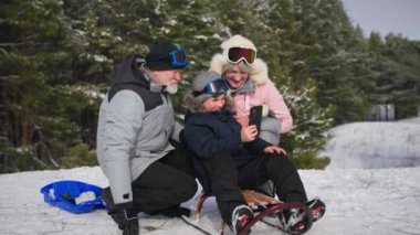 Çocuk, büyükannesiyle büyükbabasına kızağın üstünde otururken video gösteriyor. Büyükannesi ve büyükbabası ormanda torunuyla eğleniyor.