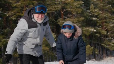 Torun büyükbabaya beşlik çakıyor ve sonra büyükannemin kızakla kaymasına yardım ediyor. Gri saçlı yaşlı kadın ormanda açık hava aktiviteleri sırasında cep telefonuyla video çekiyor.