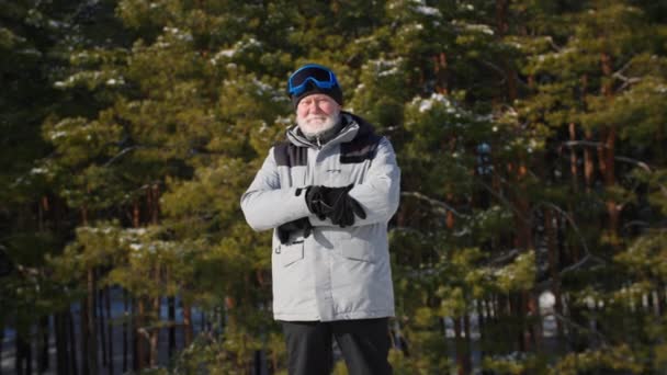 Portrét důchodce s šedým vousem zkříží ruce a dívá se do kamery na pozadí lesa, starší muž v teplých šatech a lyžařských brýlích v zimě — Stock video