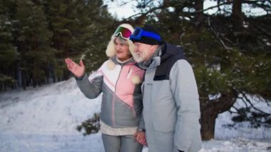 Birlikte mutlu, sıcak kıyafetler ve kayak gözlüğü takan yaşlı bir çift, karlı ormanda el ele yürürken iletişim kuruyor.