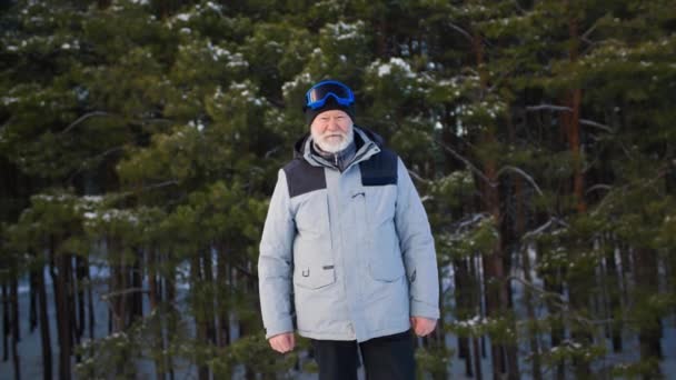 Porträt eines Rentners mit grauem Bart, der in Triumphgeste die Fäuste hebt und in die Kamera auf dem Hintergrund des Waldes blickt, eines älteren Mannes in warmer Kleidung und Skibrille im Winter — Stockvideo