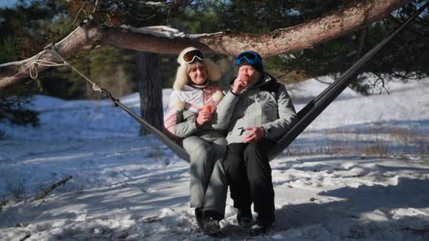 Důchodci pijí horký čaj z pohárů sedí na houpací síti a chatu, šťastný starší pár tráví víkend v zimním lese — Stock video