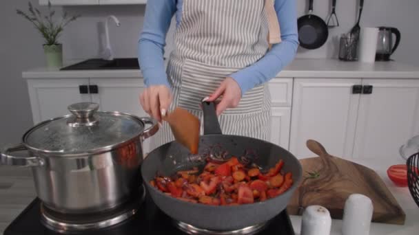 Домашнє блюдо, молода домогосподарка в фартусі використовує електричну плиту для приготування овочів на гарячій сковороді на кухні — стокове відео