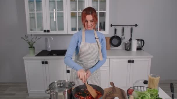 Vegetarian cuisine, young woman in an apron cooks colored vegetables in hot frying pan and moves them with spatula while standing in kitchen — Stock Video