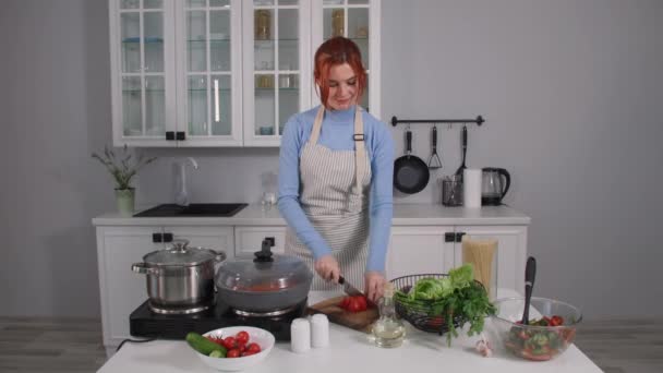 在家里做饭的过程中，一位穿着围裙的迷人女人在厨房里靠近桌子时，把西红柿切好，扔进热锅里，加入彩色蔬菜和木铲 — 图库视频影像